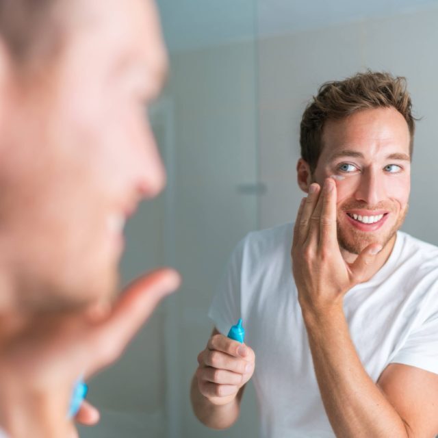 Smiling young man putting face cream under eyes to treat wrinkles or undereye eyebags, Anti-aging facial treatment. Male beauty.