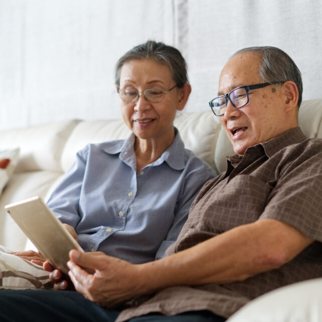Senior couple sitting on sofa in home playing tablet and relaxing together. They are smiling and enjoy to spend their time together with happiness. Happy retirement life concept.